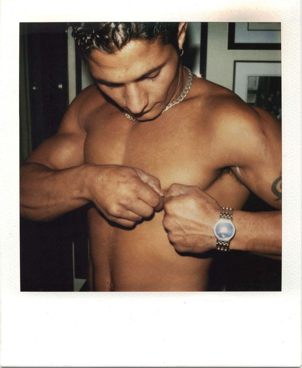 Polaroid of a shirtless muscular man wearing a chain necklace and wristwatch, looking down while fastening or adjusting jewelry with both hands indoors. Image from Michael Alago\u2019s book Polaroid Encounters