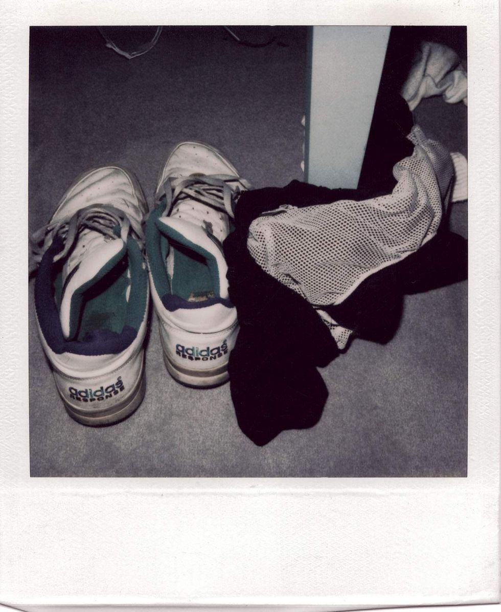 Polaroid photograph of a pair of worn white Adidas sneakers on the floor beside crumpled black shorts in a dimly lit room. Image from Michael Alago\u2019s book Polaroid Encounters