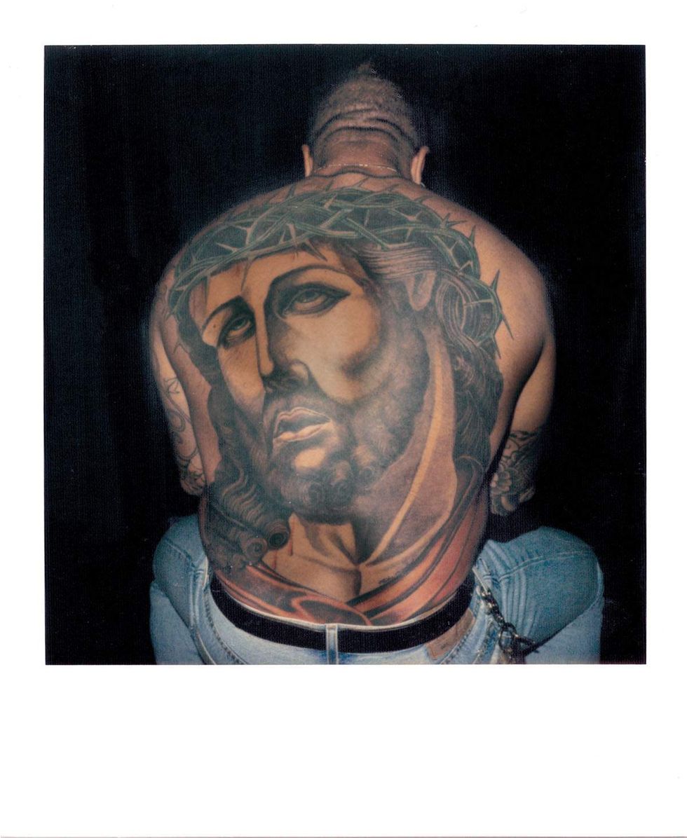 Polaroid photograph of a shirtless man turned away from the camera, revealing a large full-back tattoo portrait of Jesus wearing a crown of thorns. The man is seated in low-rise jeans against a dark background. Image from Michael Alago\u2019s book Polaroid Encounters