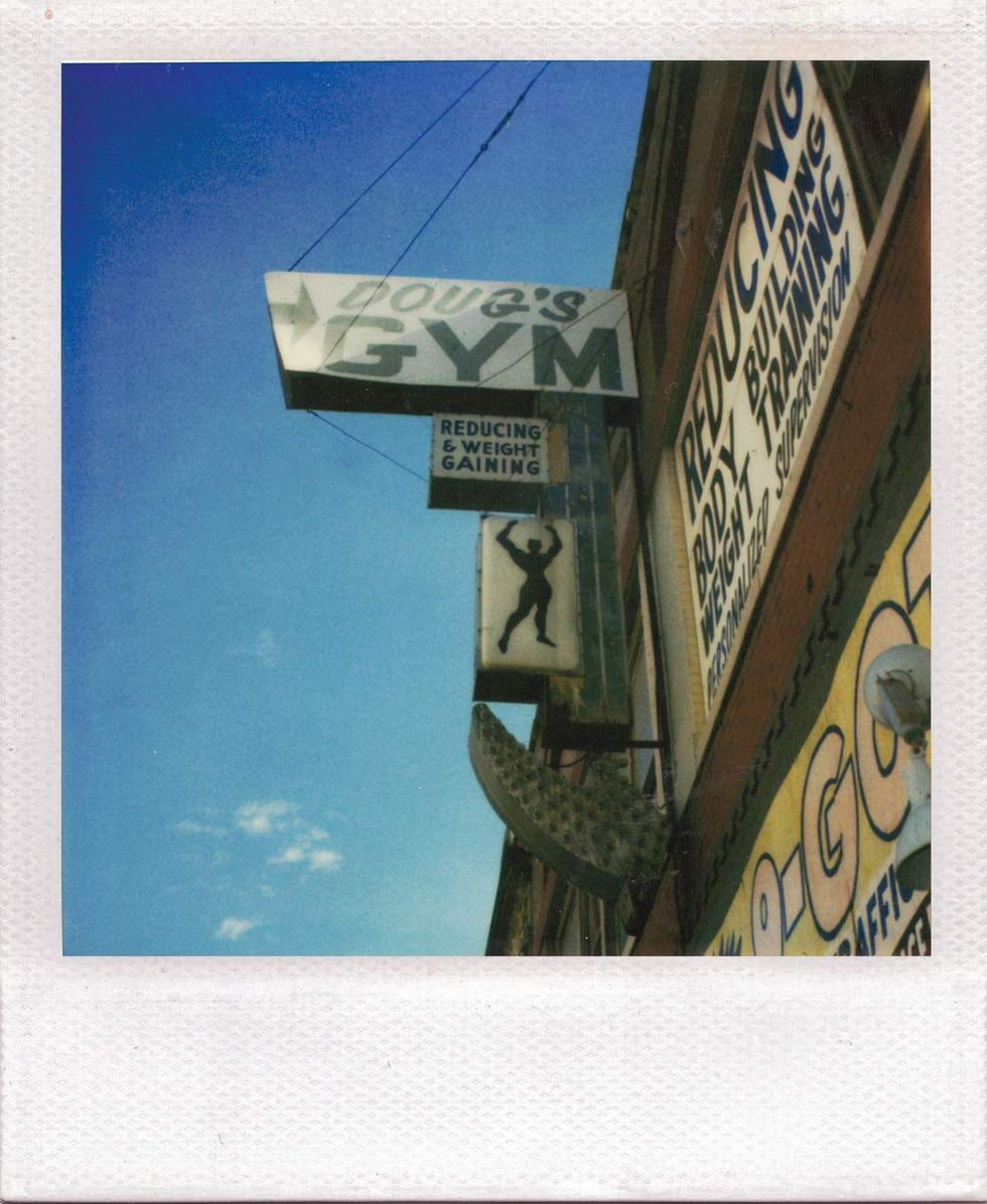 Polaroid photograph of storefront gym signs hanging above a city street, including a large sign reading \u201cDoug\u2019s Gym\u201d against a bright blue sky. Image from Michael Alago\u2019s book Polaroid Encounters