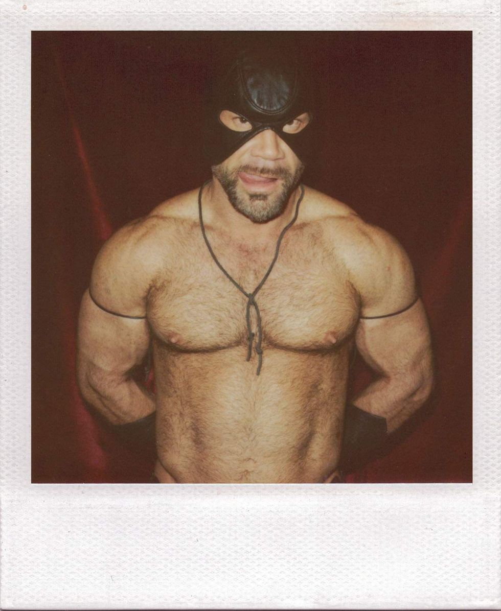 Polaroid portrait of a muscular, hairy-chested man wearing a black leather mask and thin armbands, posing with his hands behind his back against a dark red curtain backdrop. Image from Michael Alago\u2019s book Polaroid Encounters