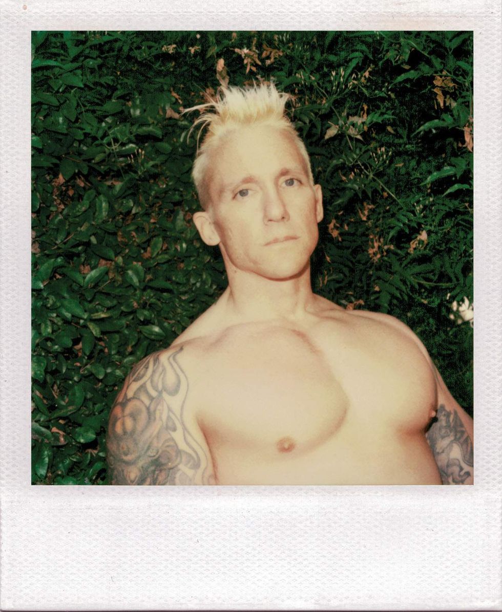 Polaroid portrait of a shirtless blond man with short spiked hair and tattooed arms posing outdoors in front of dense green foliage, looking directly at the camera. Image from Michael Alago\u2019s book Polaroid Encounters