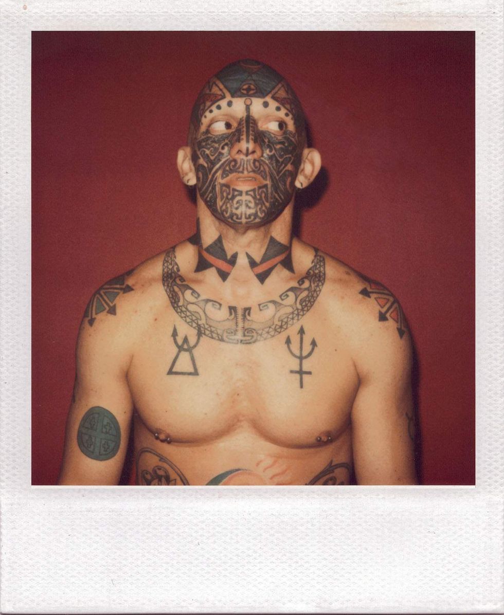 Polaroid portrait of a shirtless man covered in extensive tattoos, including intricate black facial tattoos and geometric designs across his neck and chest, posing against a deep red background while looking to the side. Image from Michael Alago\u2019s book Polaroid Encounters