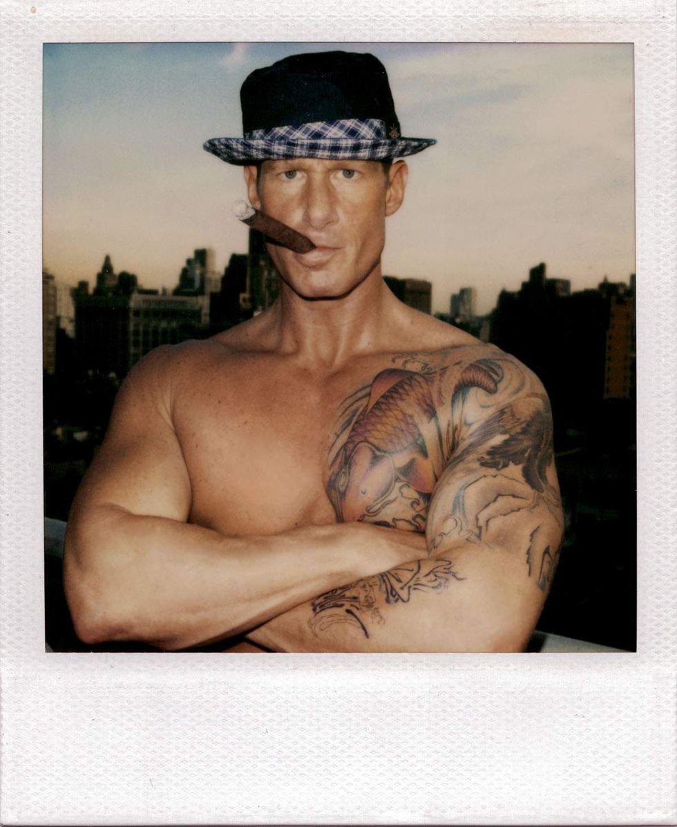 Polaroid portrait of a shirtless man with tattooed arms standing on a rooftop at dusk, wearing a dark fedora with a plaid band and holding a cigar in his mouth while crossing his arms. A city skyline appears in the background