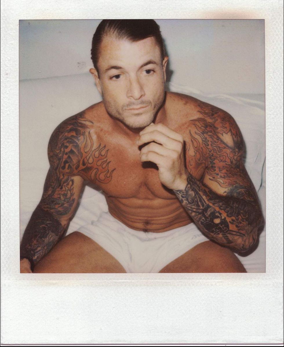Polaroid portrait of a shirtless, muscular man with slicked-back dark hair and heavily tattooed arms sitting on a bed in white briefs, resting one hand near his chin and looking off-camera. Image from Michael Alago\u2019s book Polaroid Encounters
