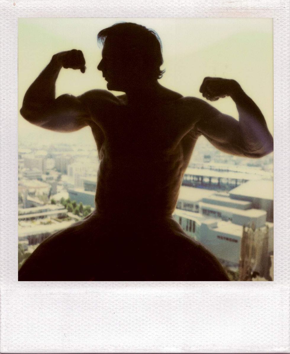 Polaroid silhouette of a muscular man flexing both arms while seated in front of a large window overlooking a sunlit cityscape. The figure appears in shadow against the bright skyline. Image from Michael Alago\u2019s book Polaroid Encounters