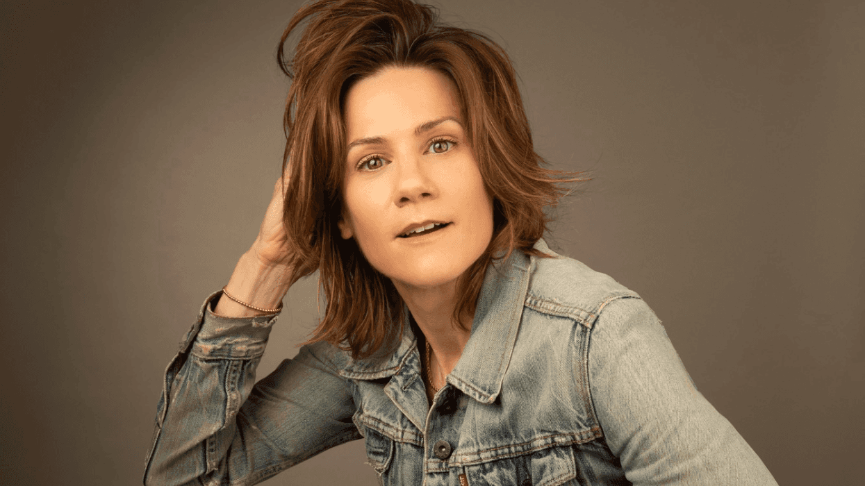 Portrait of Cameron Esposito in a jean jacket, touching her hair with her right arm.