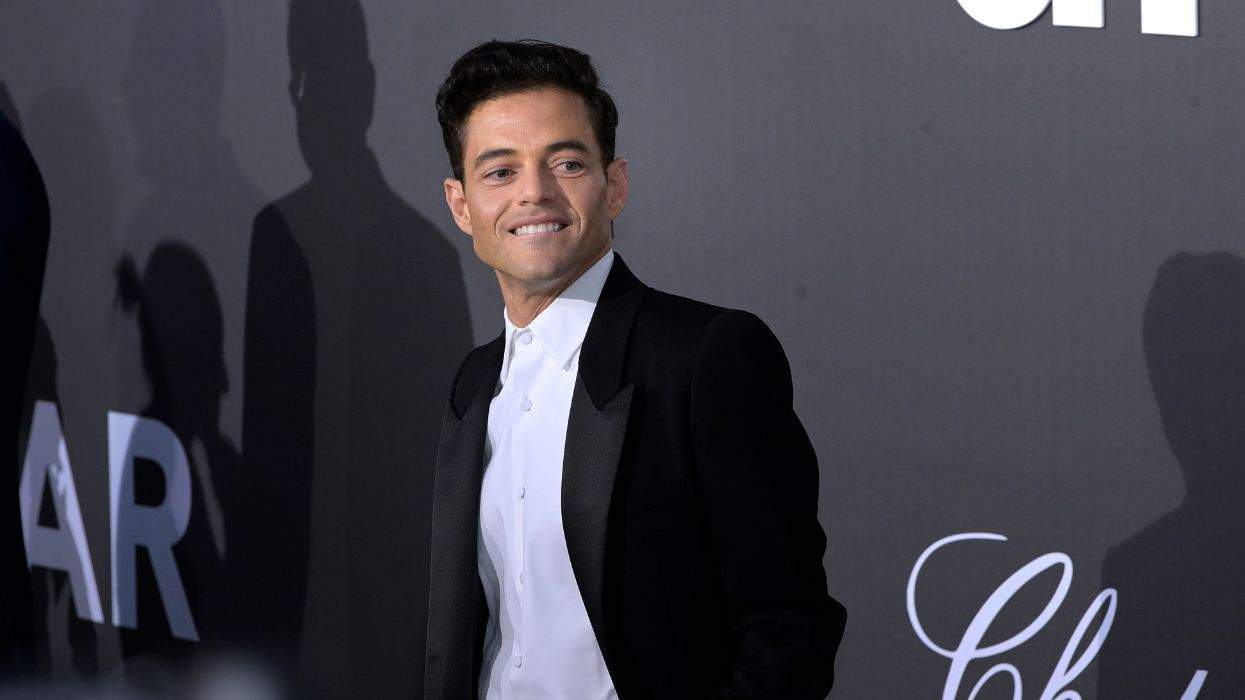 Rami Malek in a black suit smiles at a formal event with a gray backdrop