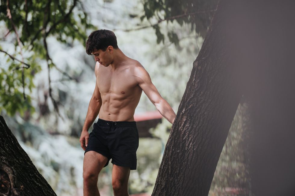 shirtless man in black shorts hiking in the woods near a lake seen between trees