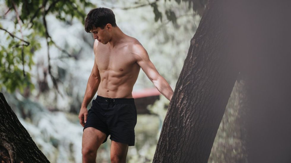 shirtless man in black shorts hiking in the woods near a lake seen between trees