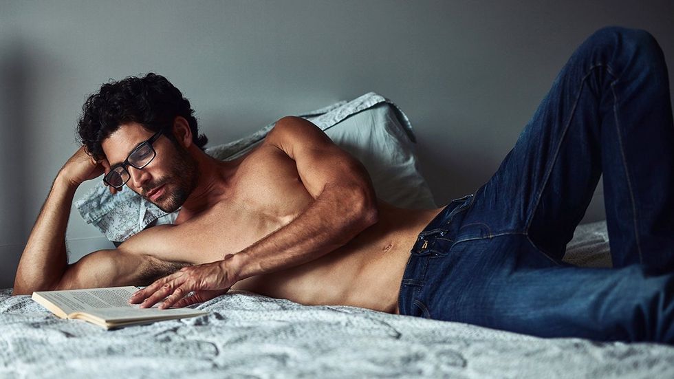 shirtless man in jeans reading book in bed