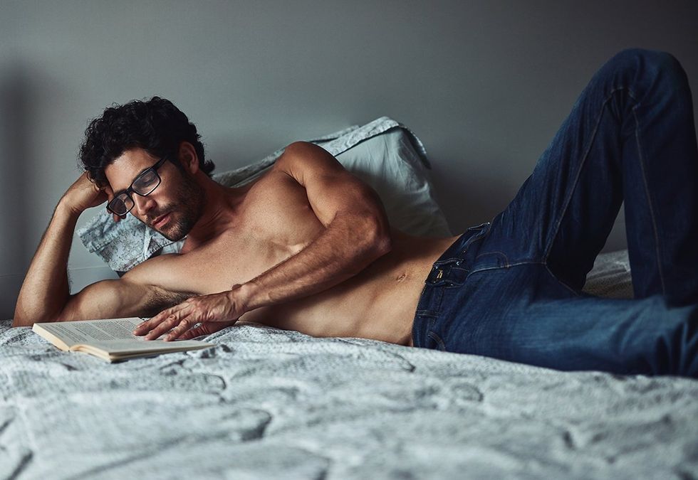 shirtless man in jeans reading book in bed