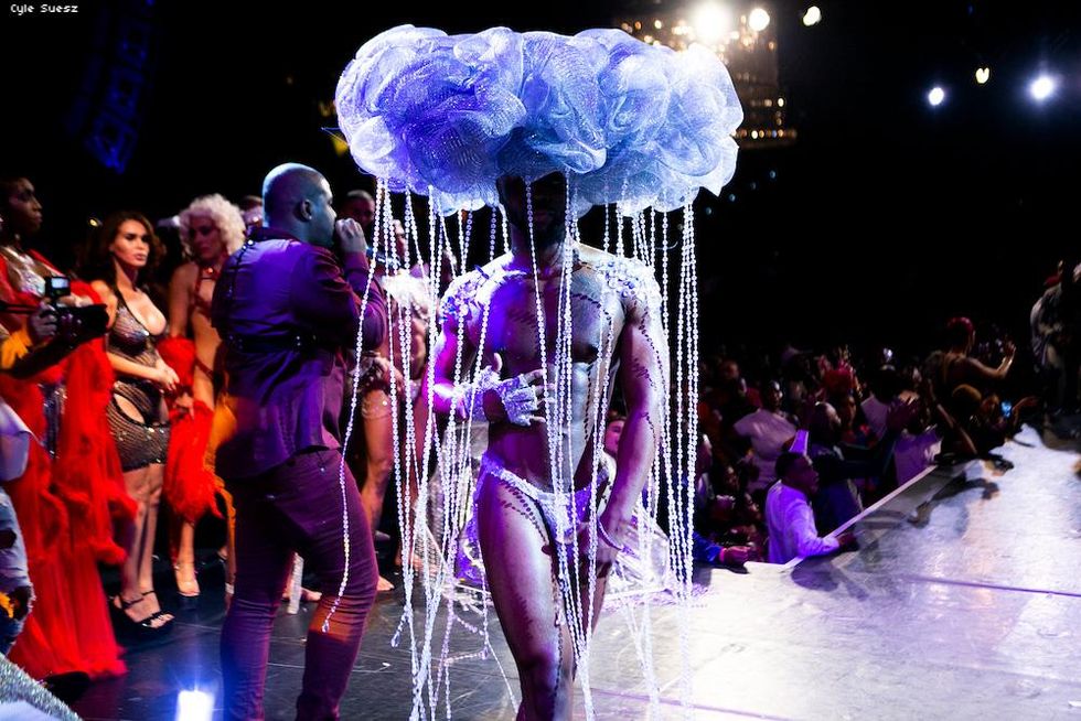 Shirtless muscular man in underwear with a headpiece that looks like a cloud that has crystals dripping from it to mock rain.