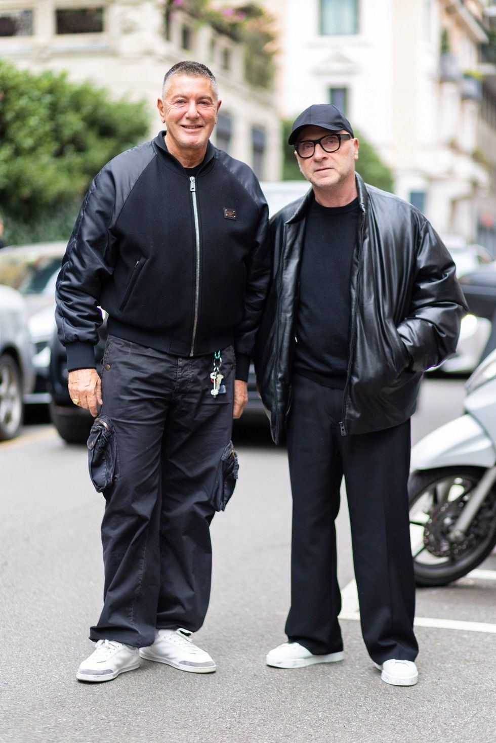 Stefano Gabbana wears a black wool and leather Dolce e Gabbana bomber, black cargo trousers, and white sneakers; and Domenico Dolce wears a black baseball hat, black glasses, a black sweater, a black leather winter jacket, black trousers, and white sneakers during Milan Fashion Week in March 2025