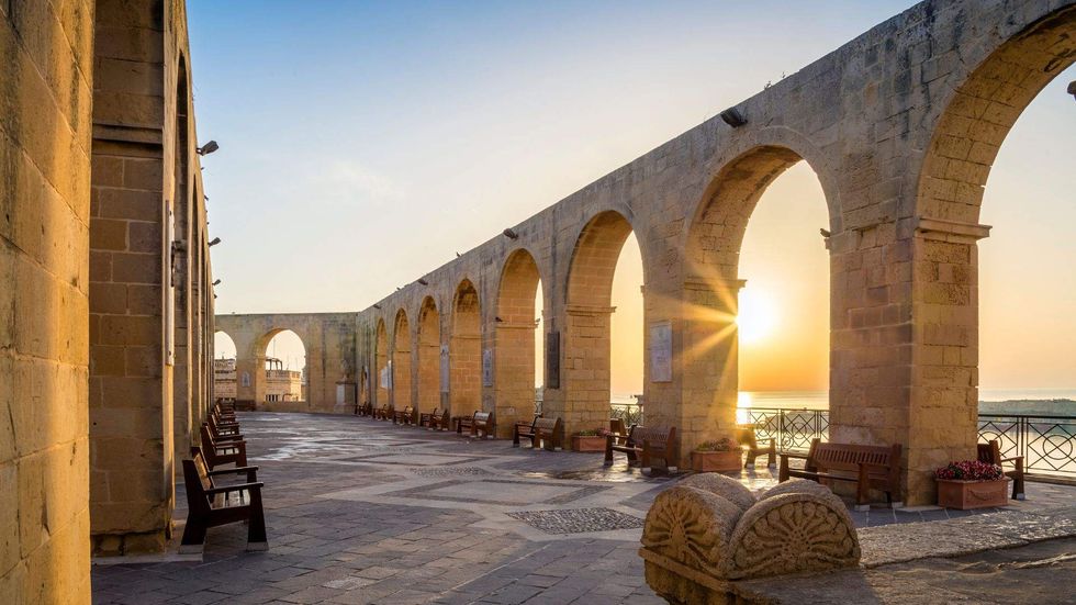 Stone arches and benches in Valletta, Malta, illuminated by sunset light overlooking the Mediterranean Sea.