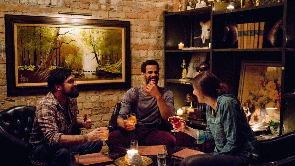 Three friends laughing and drinking in a cozy, candlelit room with art and books on shelves.