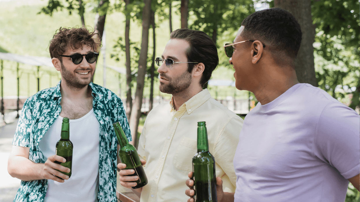 Three men wearing sunglasses chat outdoors on a sunny day, each holding a green glass beer bottle.