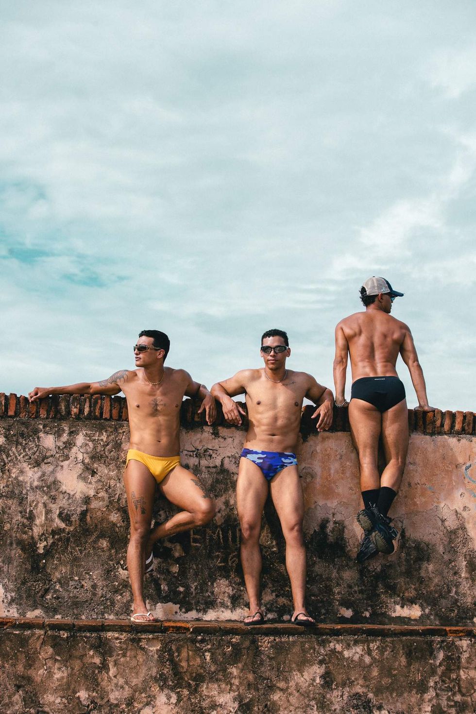 Three models wearing Sniffies swim briefs pose together on a stone wall during the Sniffies Summer Swim Collection campaign in Puerto Rico