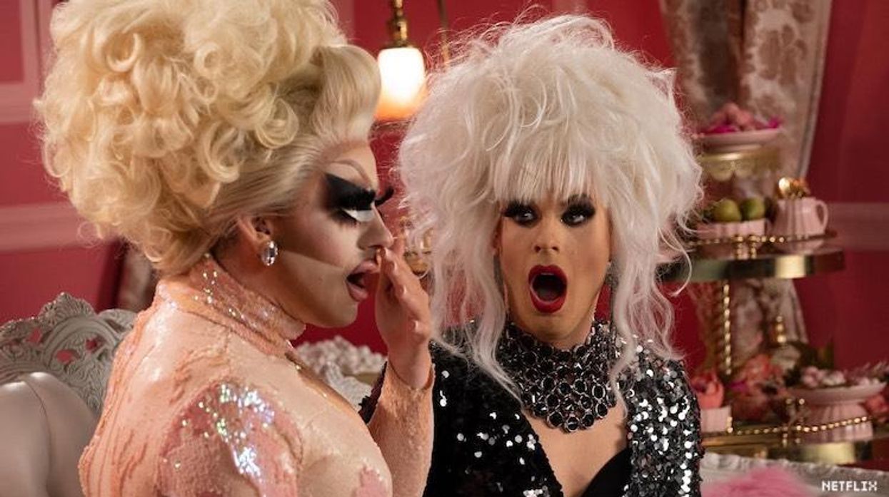Trixie and Katya in drag sitting on a couch looking surprised.