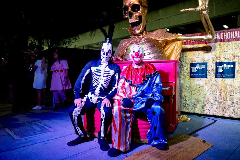 Two costumed attendees sit in front of a red piano beneath a giant gold skeleton.