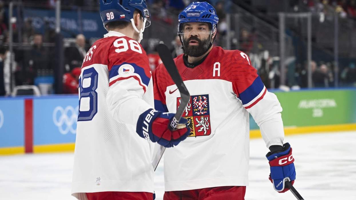Two men in red and white jerseys and blue helmets stand on a hockey rink and talk to one another, with hockey sticks in hand.
