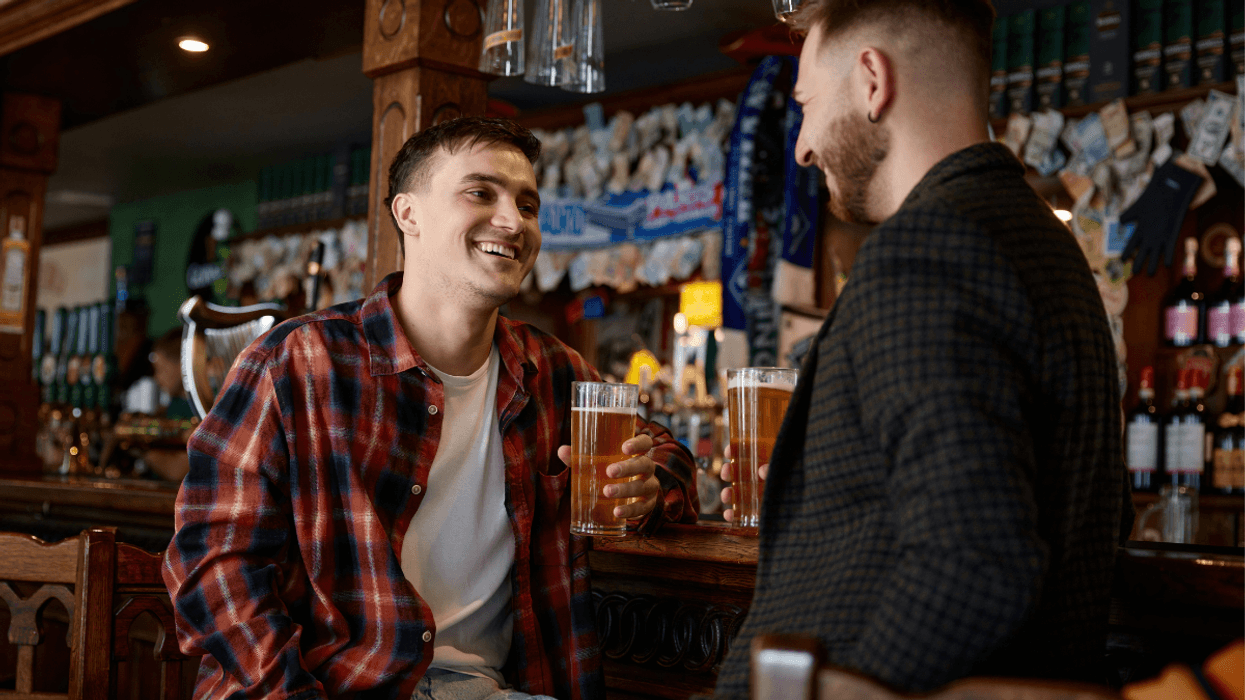 Two men smiling and talking over beers while seated at a bar.