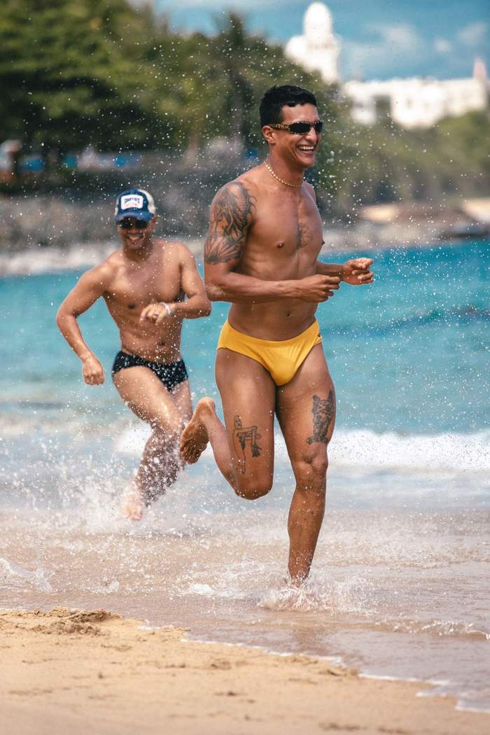 Two models wearing Sniffies swim briefs share a playful moment while running through the ocean during the Sniffies Summer Swim Collection campaign in Puerto Rico