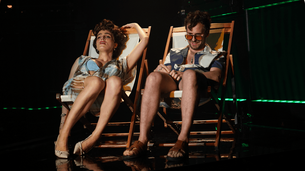 Two people sitting in fold-out chairs on stage in beach attire.