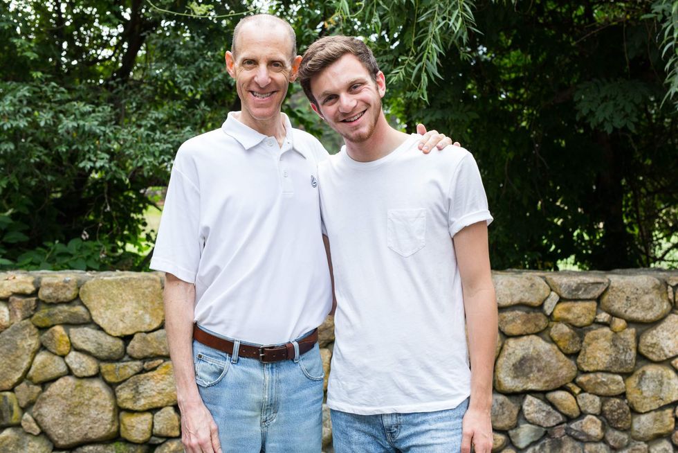 Two people smiling in front of a stone wall and trees, wearing white shirts and jeans.