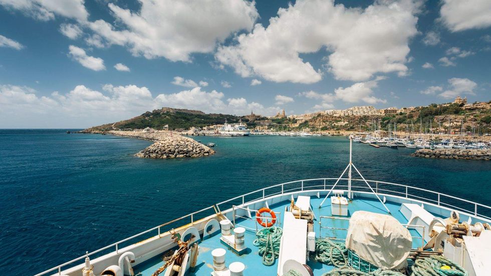 View from a ferry approaching Gozo, Malta, with turquoise water, rocky coastline, boats, and hillside buildings under a blue sky