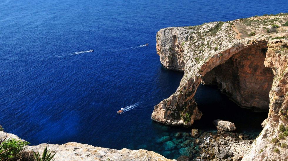 View of the Blue Grotto in Malta with rocky sea cliffs, deep blue water, and small boats passing by