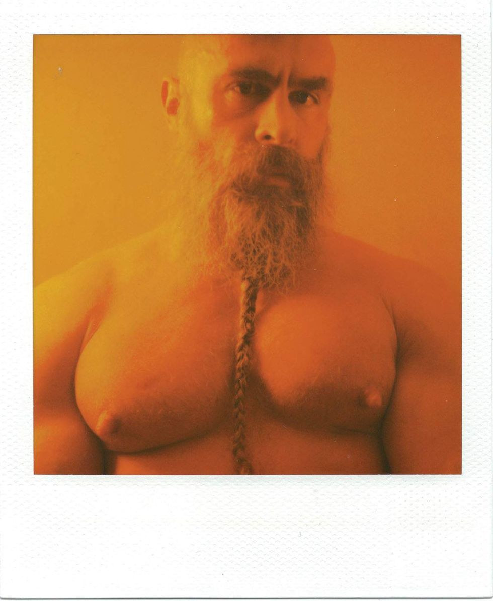 Warm-toned Polaroid portrait of a shirtless man with a long braided beard and shaved head, staring directly at the camera against an orange background. Image from Michael Alago\u2019s book Polaroid Encounters