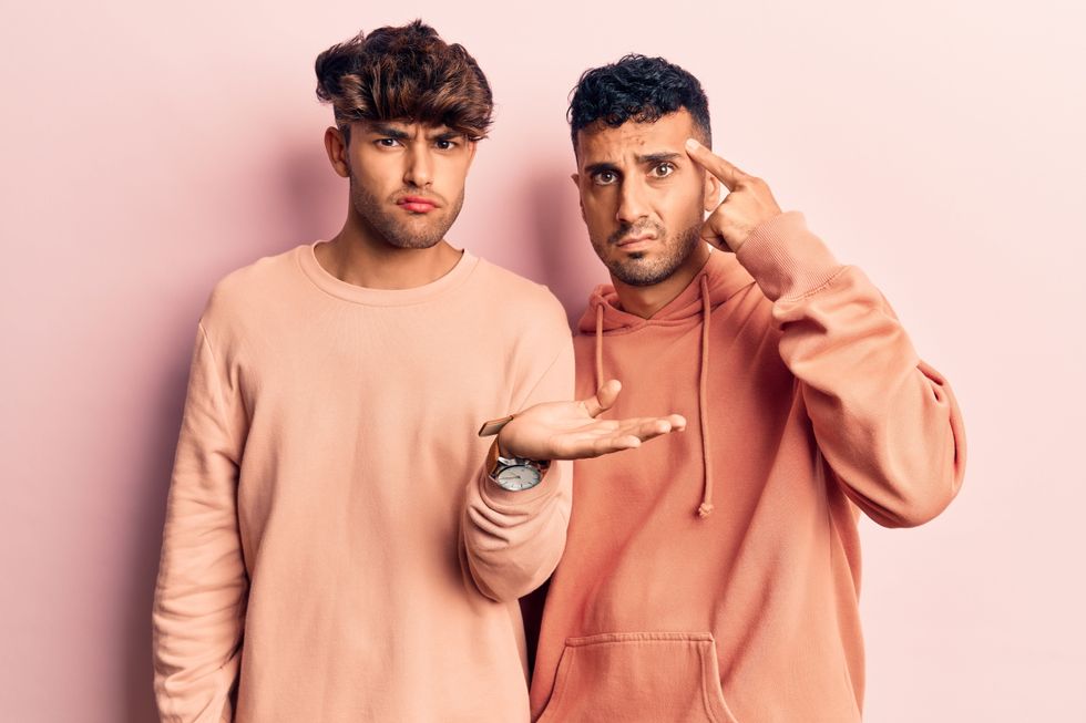 Young gay couple wearing casual clothes confused and annoyed with open palm showing copy space and pointing finger to forehead. think about it.