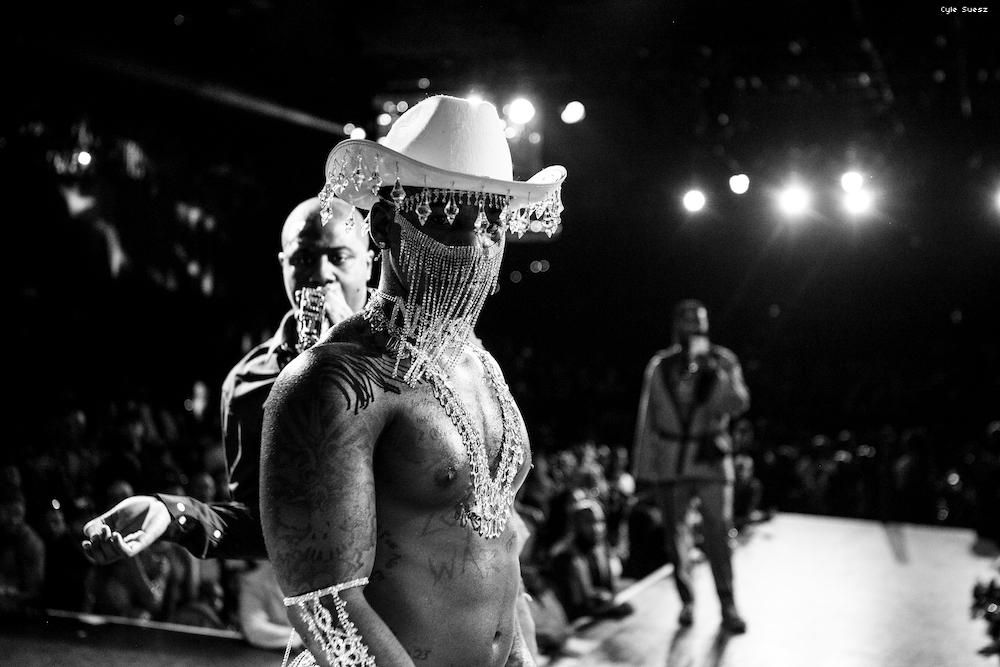 A black and white photo of a muscular, tattooed black man wearing a jeweled cowboy hat.