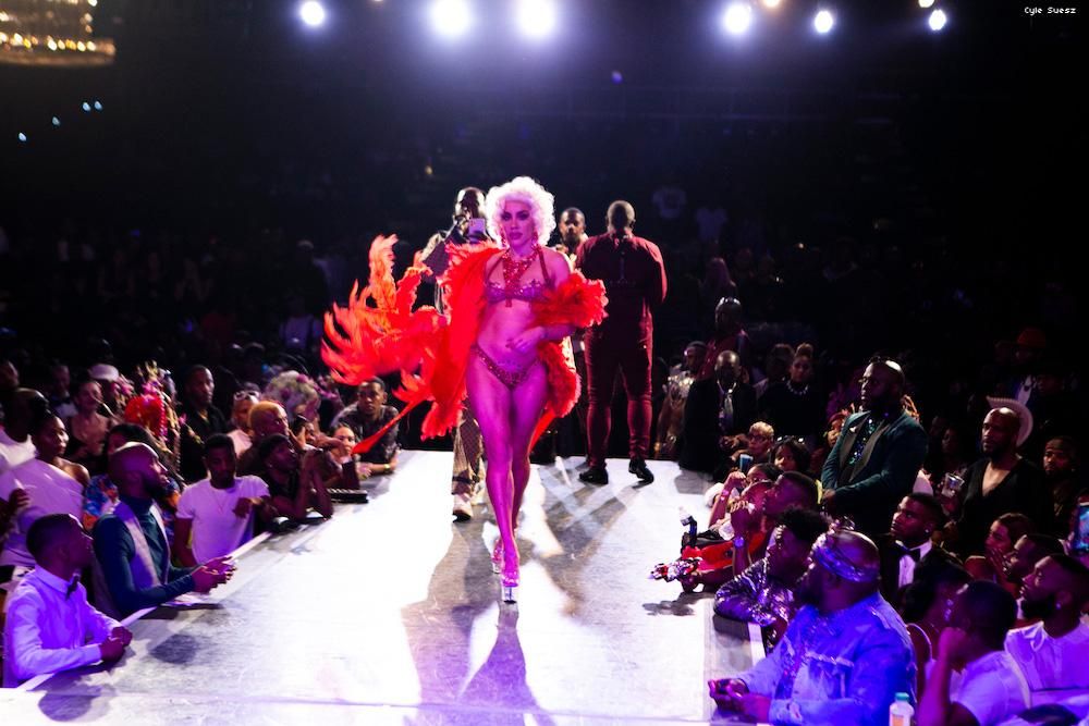 Woman in a red boa, thong and bra with blonde hair, walking down the runway.