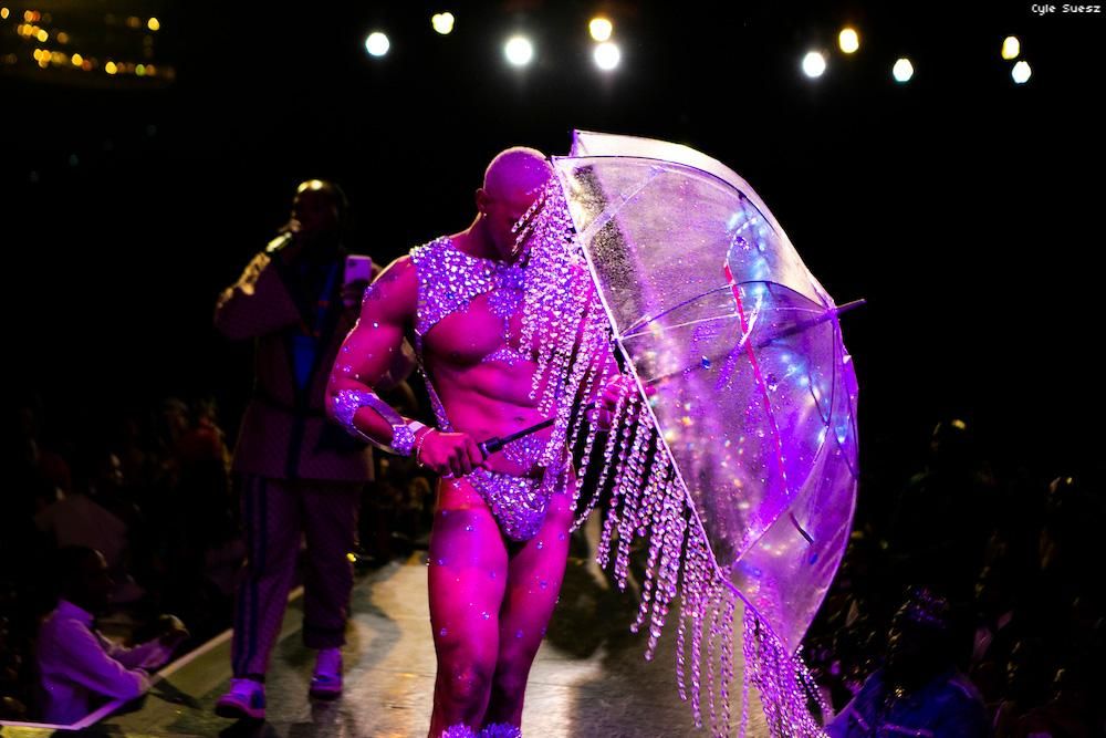 Man in a bejeweled thong emerging from under a clear umbrella dripping with crystals.