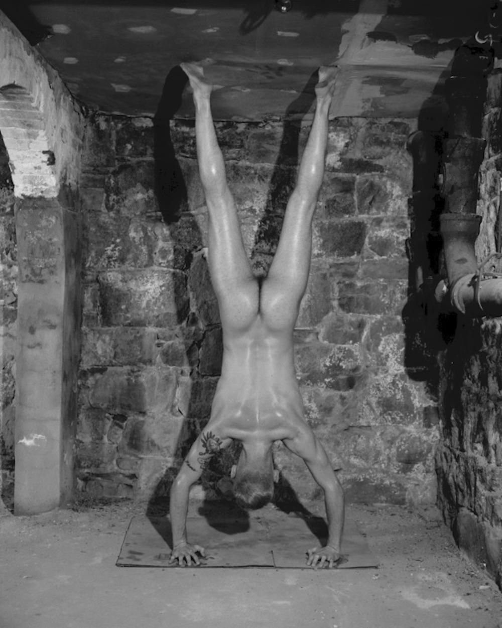 A nude male model posing in a handstand, from behind.